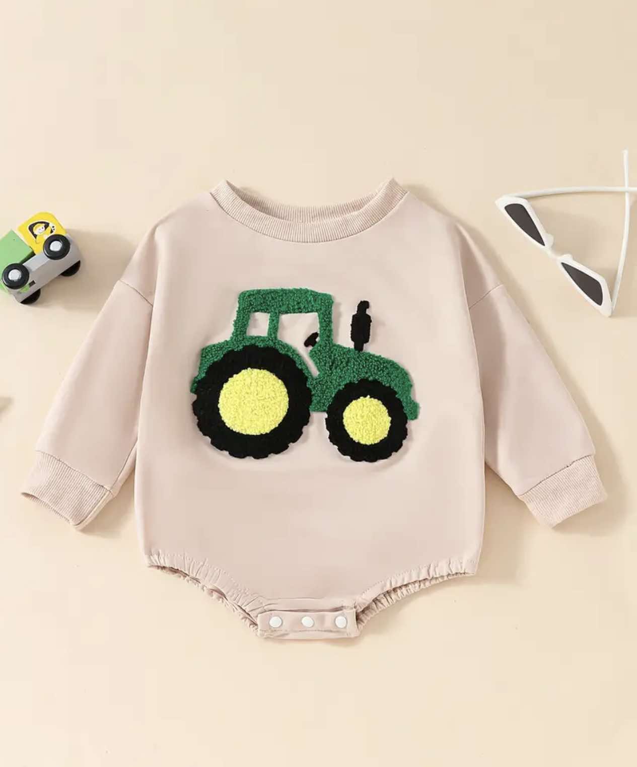 Beige long-sleeve baby onesie with an embroidered green tractor on the chest, displayed flat on a wooden surface.