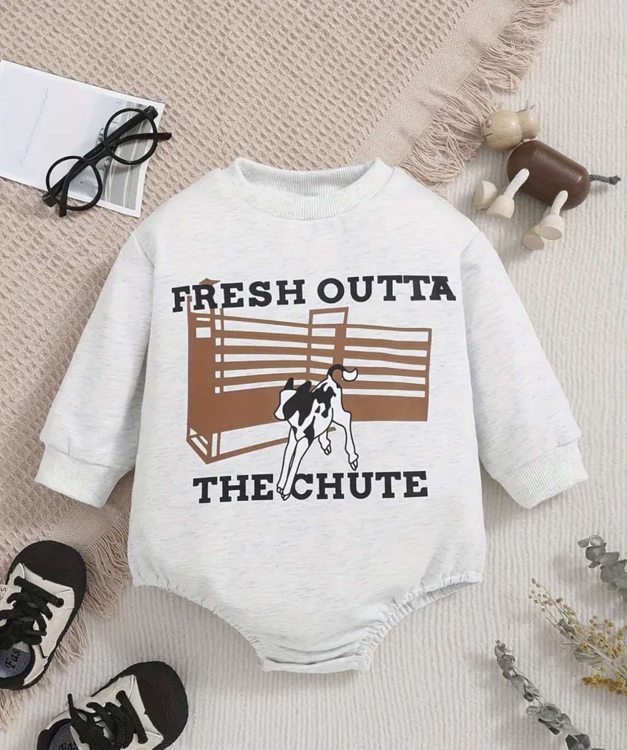 Grey oversized baby romper with 'Fresh Out of the Chute' text and cattle print, displayed flat on a wooden backdrop