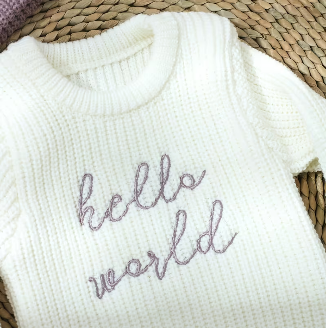 Little Wrangler Knit Romper Set from House of Hide in white ribbed knit with purple embroidered hello world text being displayed in close-up on woven straw mat