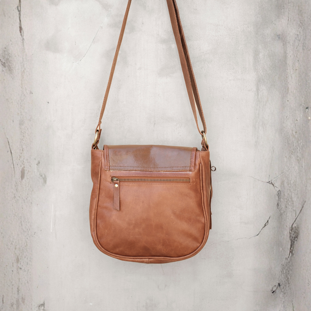 Meadow Crossbody Bag from House of Hide in brown leather with front zippered pocket and matching shoulder strap being displayed hanging against textured gray concrete wall