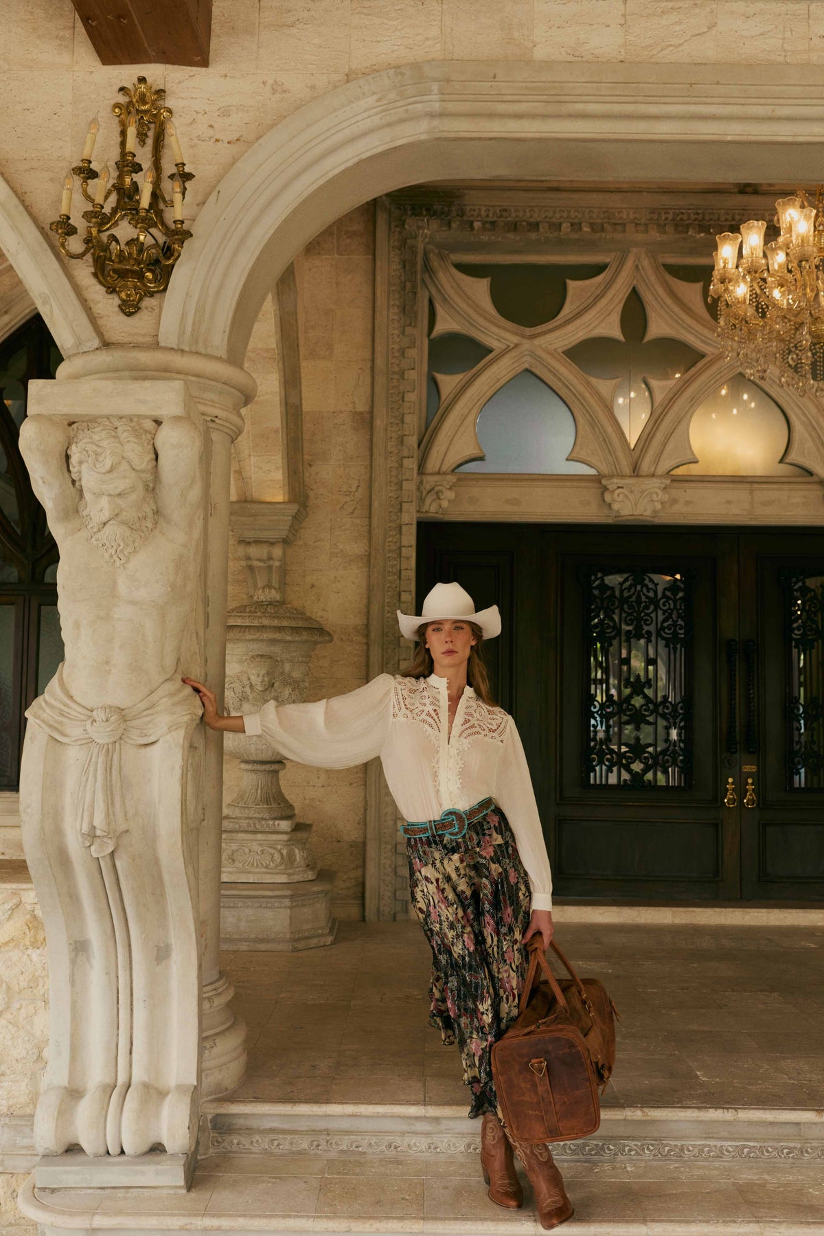Model showcasing House of Hide women's western fashion in a white shirt and cowboy hat, standing in an elegant interior with decorative columns and chandeliers