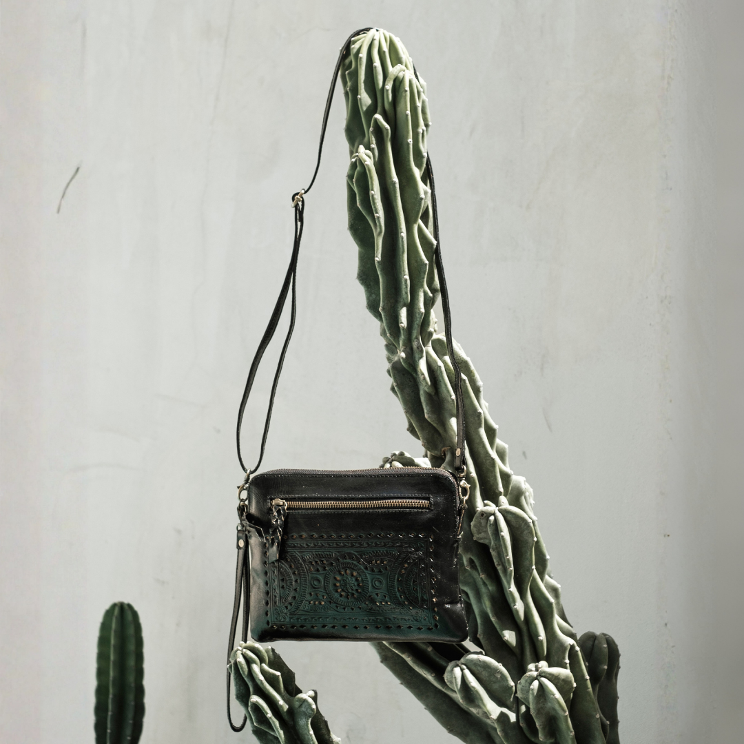 Nightshade Crossbody Bag from House of Hide in dark green leather with embossed lettering being displayed against tall green cactus and light gray textured wall