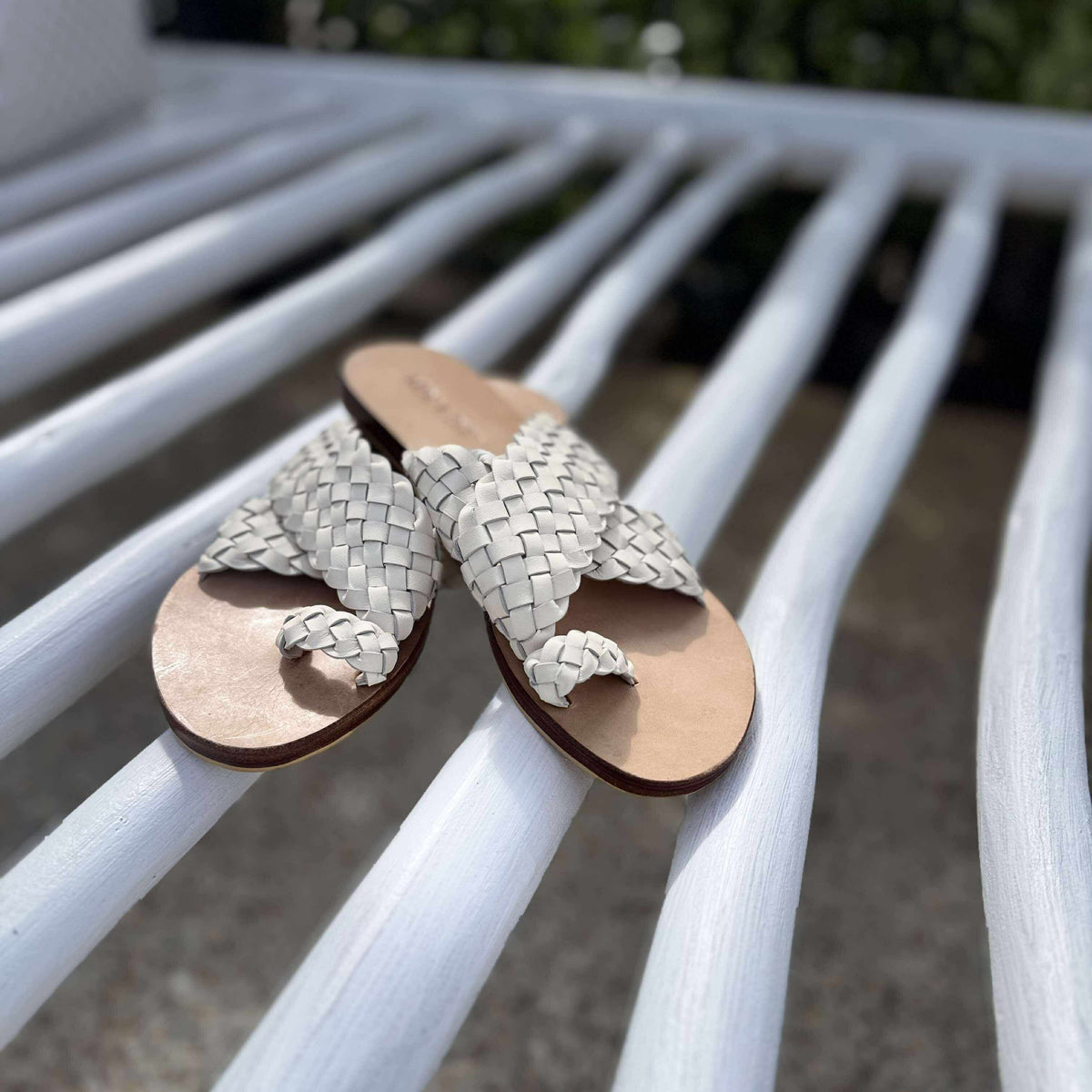 A pair of woven leather slide sandals with tan footbeds resting on the white slats of an outdoor bench.