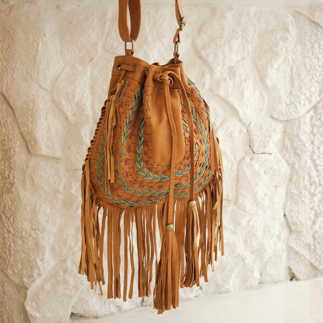 Sedona Drawstring Bag from House of Hide in tan leather with fringe detail, turquoise and orange embroidered detailing, and drawstring closure being displayed hanging against white textured stone wall