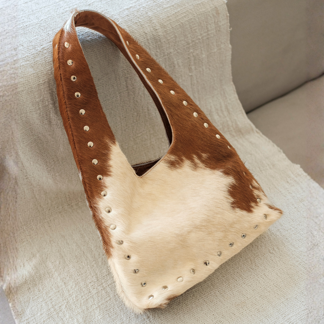 Wrangler Cowhide Bag from House of Hide in brown and cream cowhide leather with stud detailing being displayed on white couch with white textile backdrop