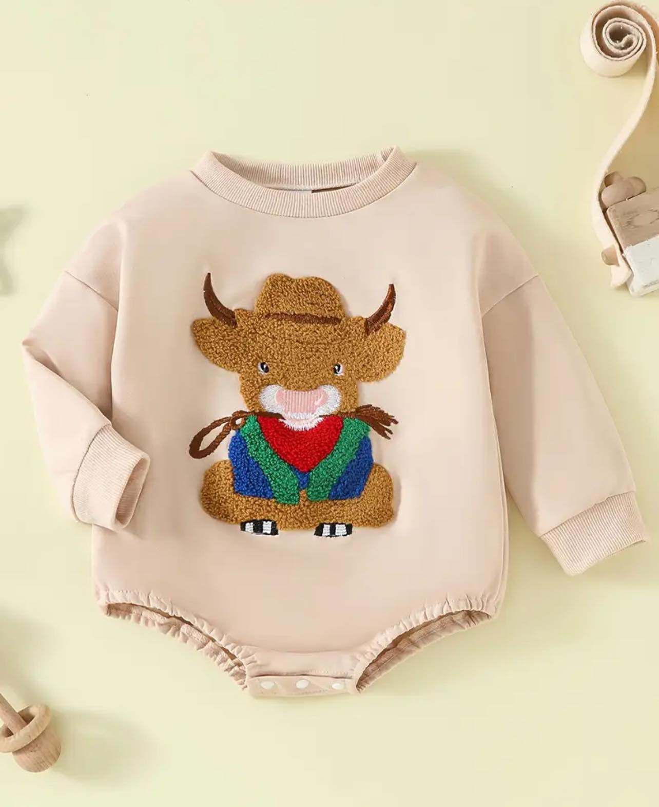House of Hide Behind the Bull romper displayed on a rustic wooden backdrop, highlighting its soft fabric, neutral beige tone, and embroidered bull motif—perfect for country kids.