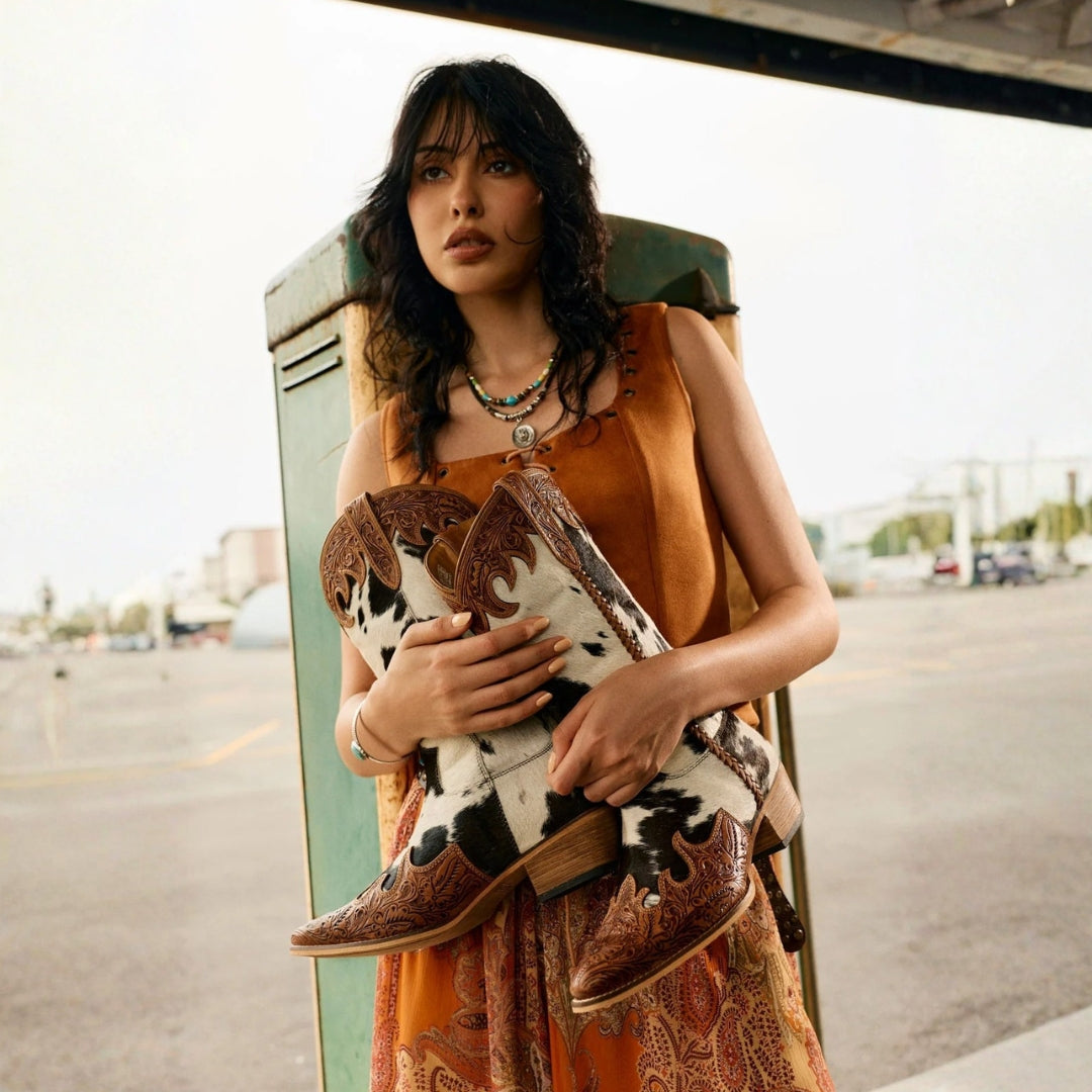 Model holding Adelaide cowhide cowboy boots with honey-toned floral tooling at the toes and tops.