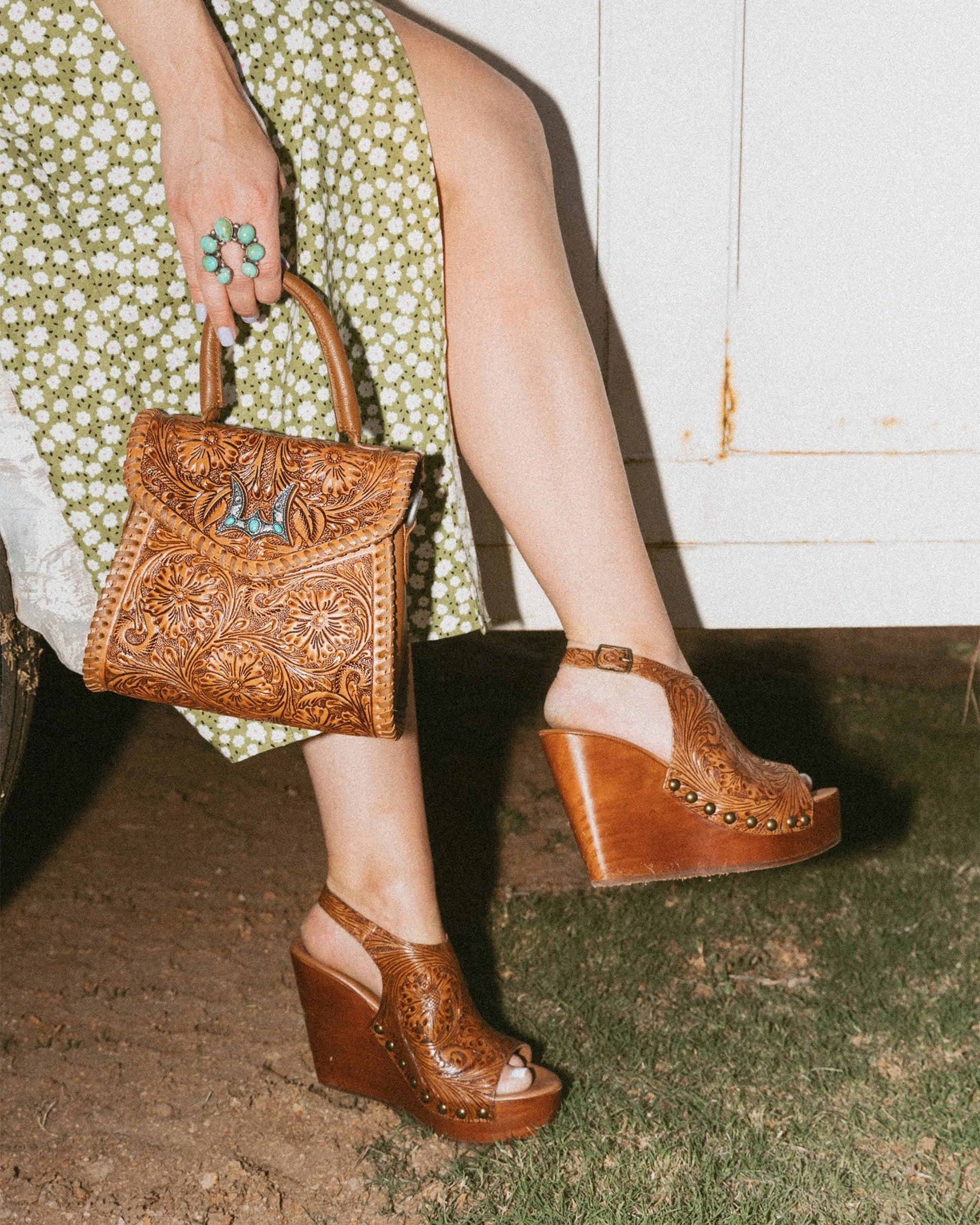 Amelia Sandals from House of Hide in brown leather with intricate tooled design and metal stud details, wooden wedge heel, being worn with matching brown tooled leather handbag against outdoor white architectural backdrop
