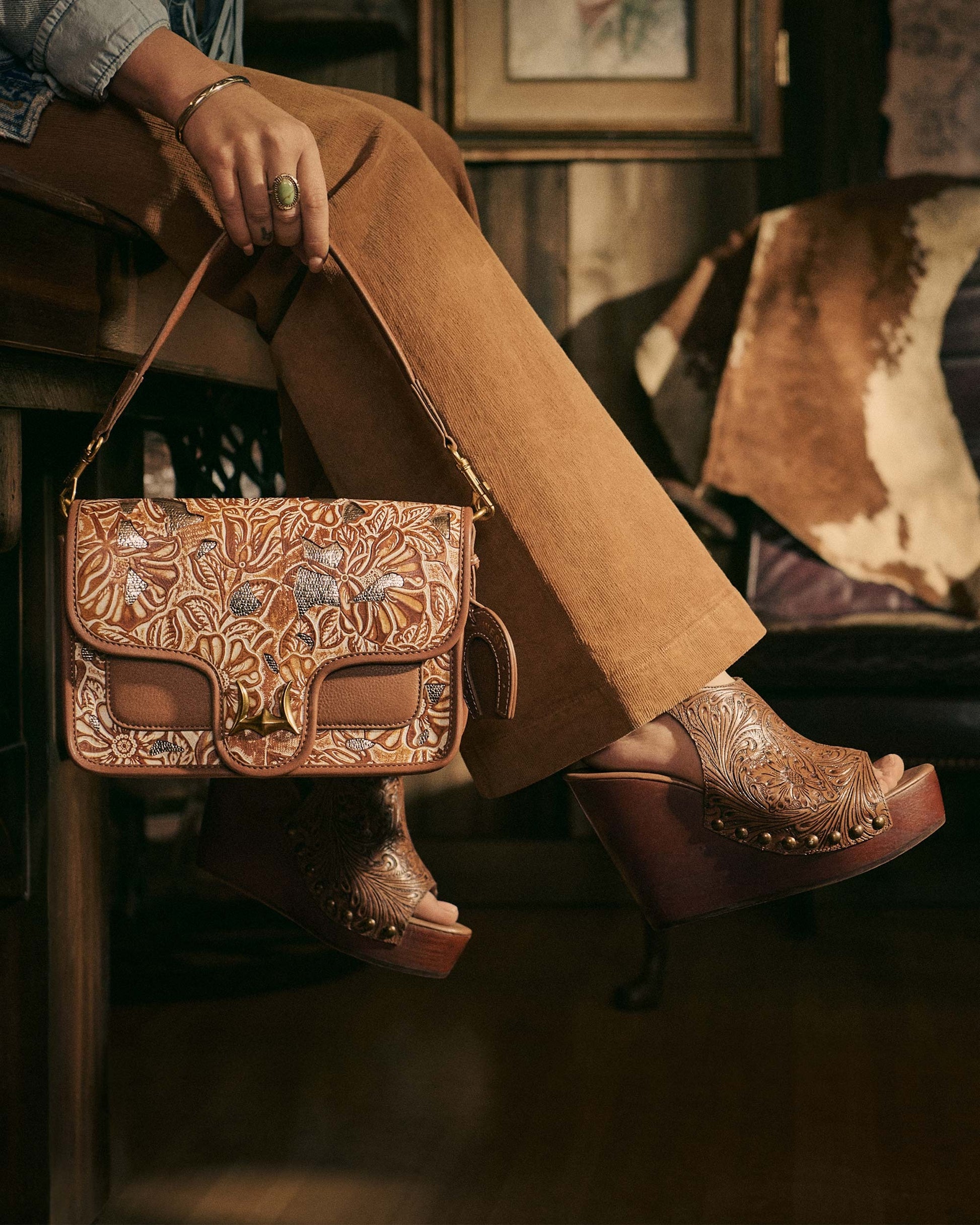 Amelia Sandals from House of Hide in brown leather with intricate tooled design and metal stud details, wooden platform wedge, being worn with matching brown tooled leather shoulder bag with turquoise stone accent in rustic interior setting