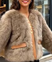 A woman wearing a fluffy taupe-brown faux fur jacket with tan leather-like trim and front buttons, photographed in a real storefront setting.