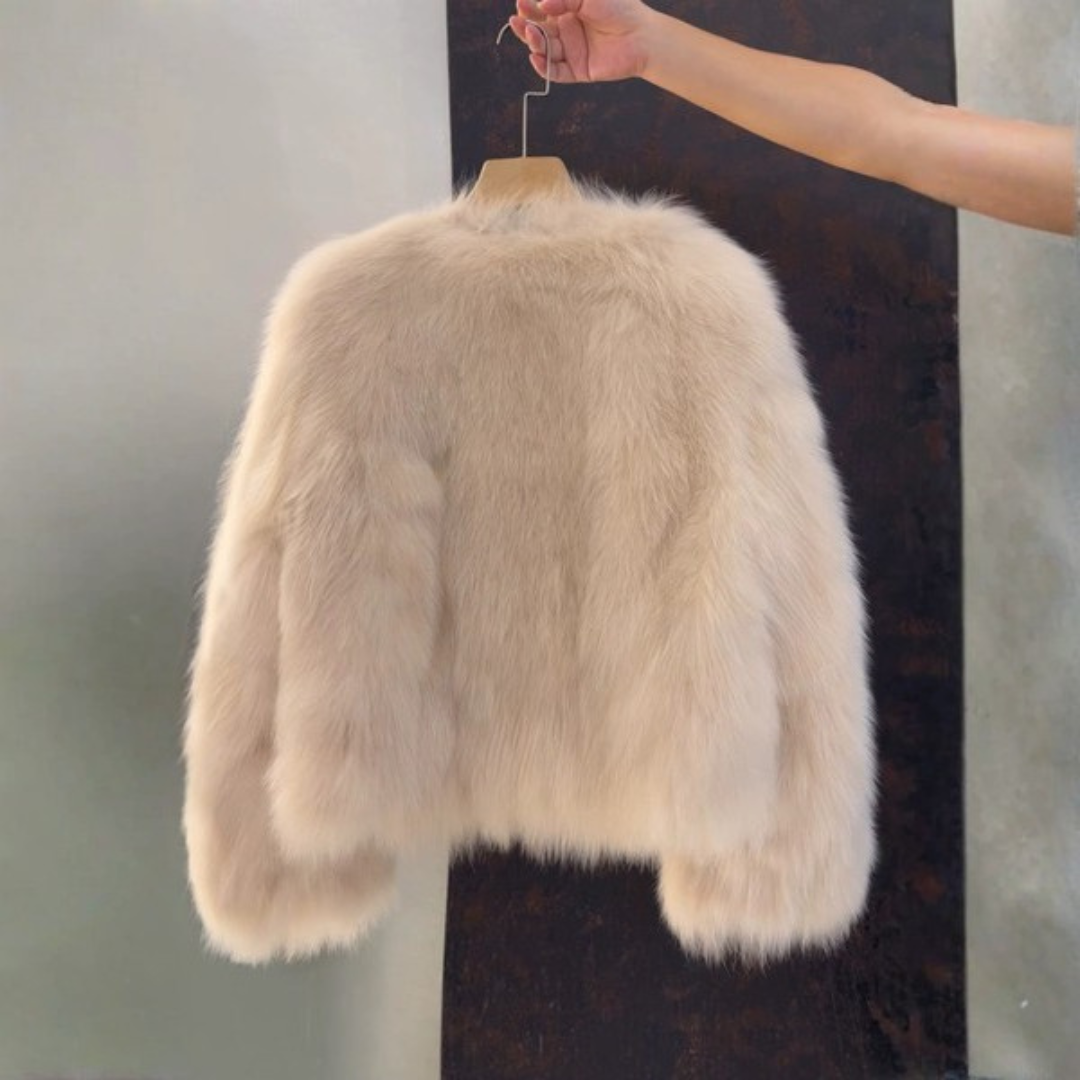 Bailey Jacket from House of Hide in plush cream faux fur with gold button closures, vegan leather placket and pocket trim, full voluminous sleeves, cropped boxy silhouette being held on wooden hanger by model's hand against split gray and dark brown background