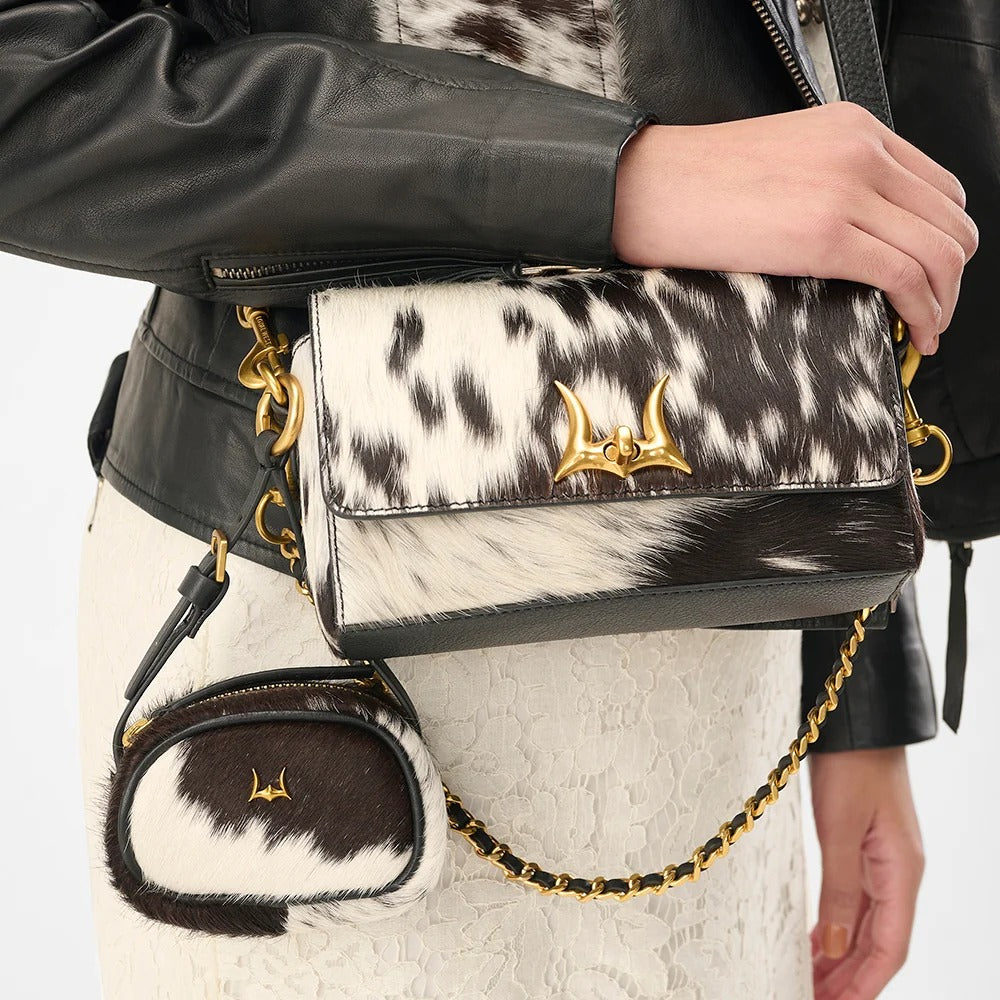 House of Hide genuine leather black and white cow print handbag with gold chain strap carried by a model wearing a black leather jacket