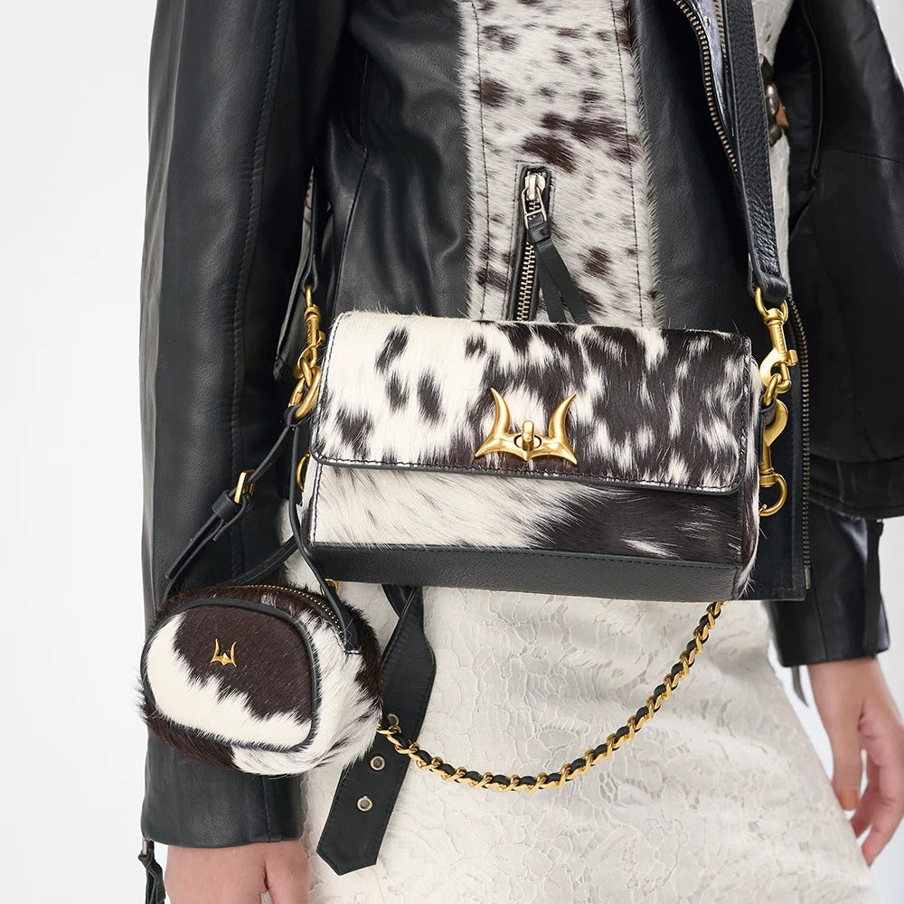 House of Hide women's western fashion featuring a genuine leather black and white cow print handbag with gold chain strap worn by a model in a black leather jacket