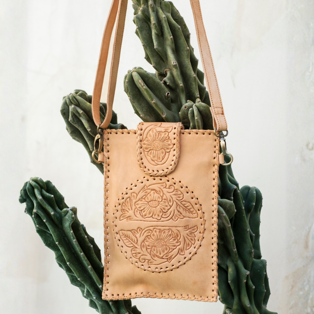 Blossom Small Crossbody Bag from House of Hide in tan leather with floral tooling and crossbody strap being displayed against light textured wall with green cactus