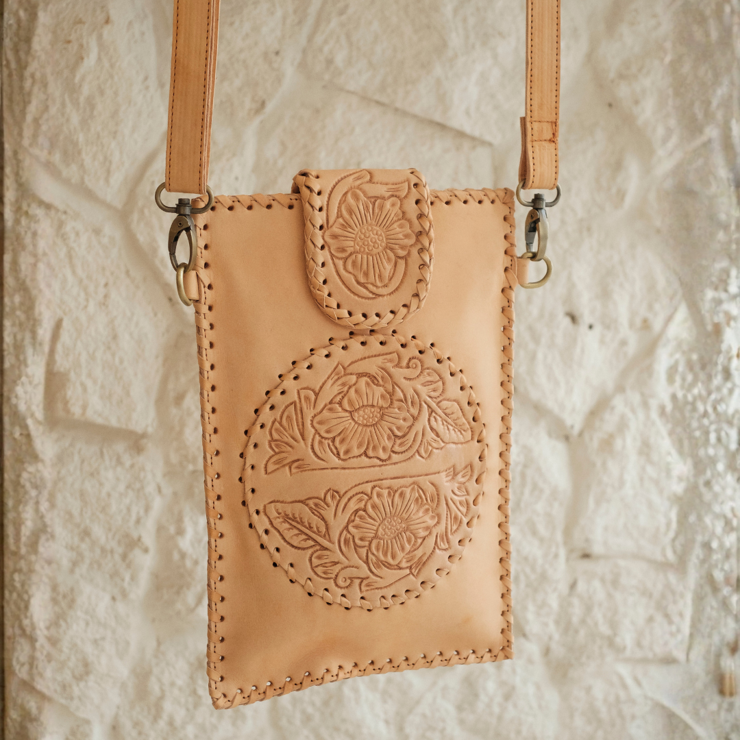 Blossom Small Crossbody Bag from House of Hide in light tan leather with floral tooling and decorative stitching being displayed against textured white stone wall