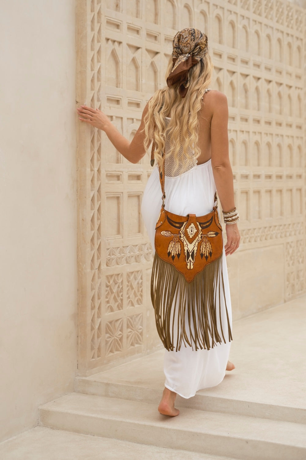 A woman with long blonde hair is seen from behind, wearing a white dress and a patterned headwrap while carrying a brown fringe crossbody bag with gold-toned embellishments, standing on stone steps against a beige geometric wall.
