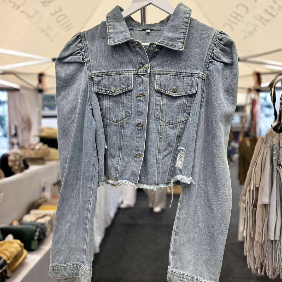 A light-wash blue denim jacket with puff sleeves and distressed details, hanging on a white hanger in what appears to be a market or boutique setting.