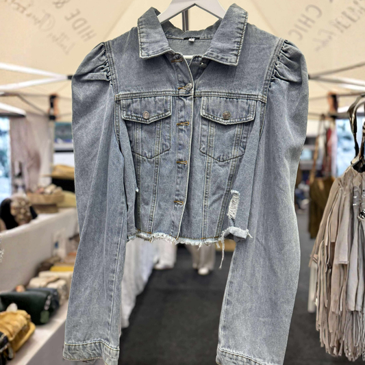 A light-wash blue denim jacket with puff sleeves and distressed details, hanging on a white hanger in what appears to be a market or boutique setting.