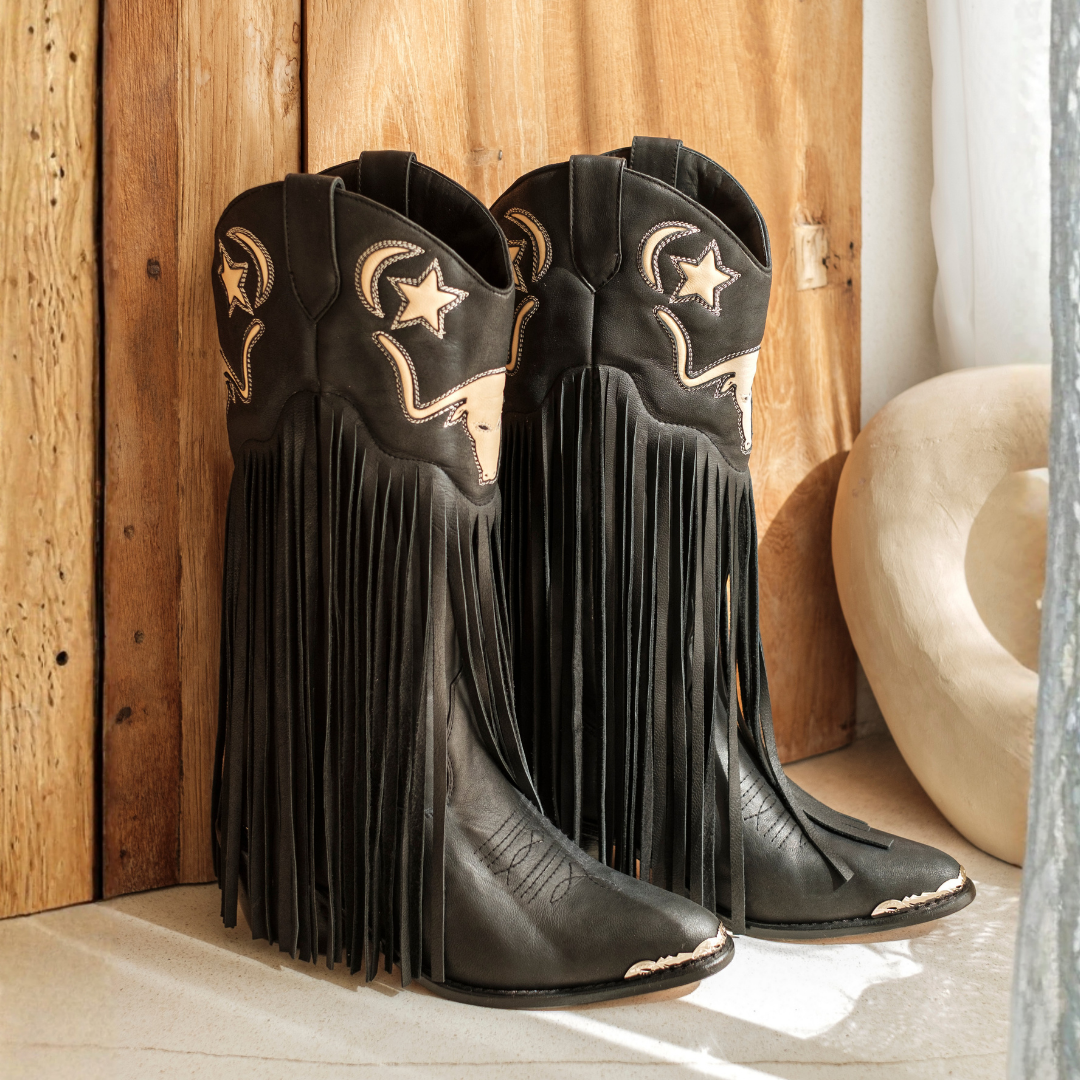 A pair of black leather cowboy boots with long fringe, embroidered moon and star designs, and light-colored decorative accents, standing on a light surface against a rustic wooden plank background.