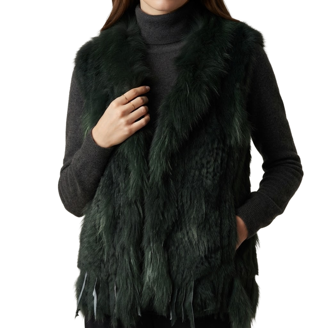 A woman wears Cottontail Fur Vest from House of Hide in bold deep forest green 100% real rabbit fur with plush raccoon fur collar, fringed hem detail, single button closure, lightweight sleeveless silhouette, 62-63cm length being styled in editorial fashion photography over jumper as edgy winter layering piece