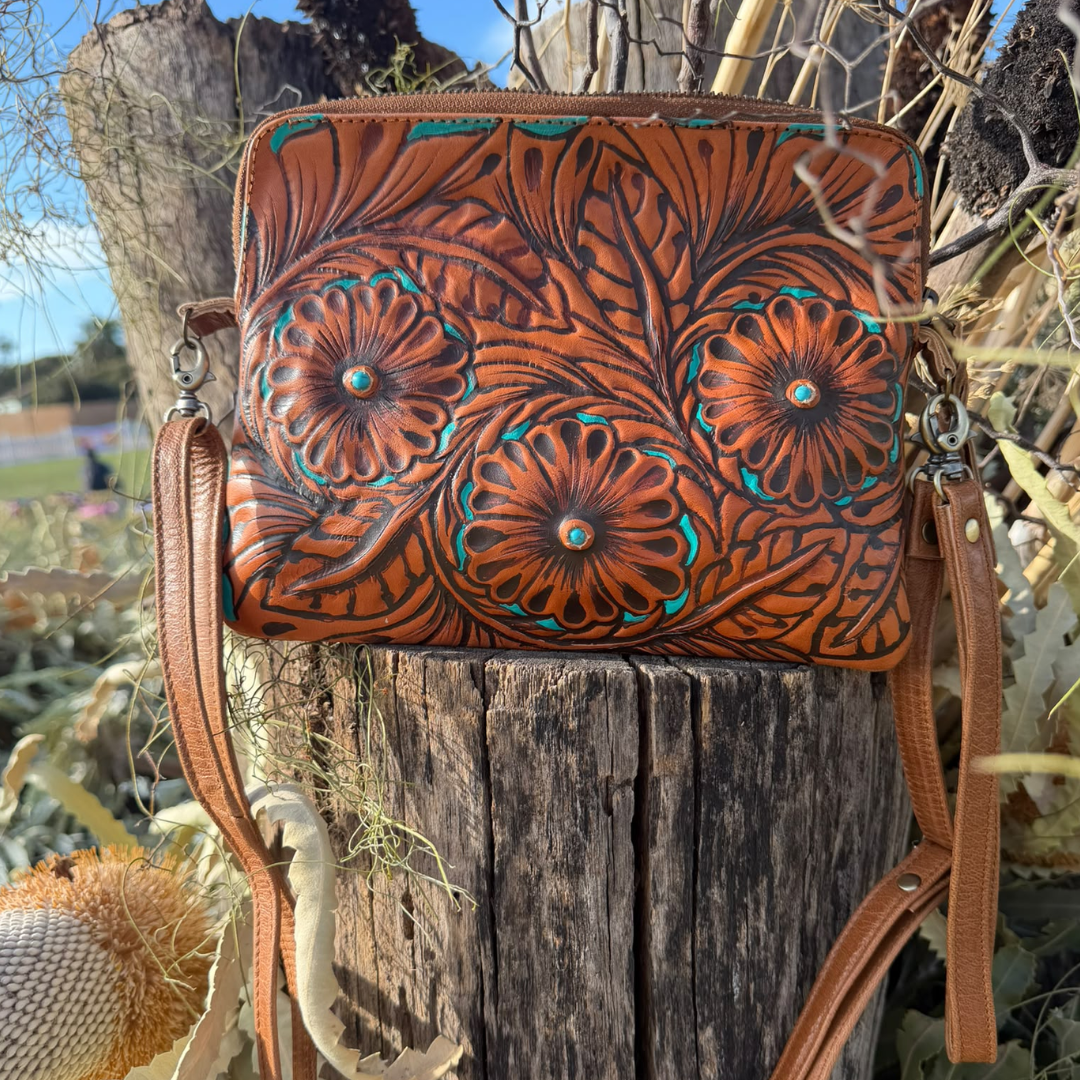 A tooled leather crossbody bag with floral designs, displayed on a rustic wooden tree stump outdoors.