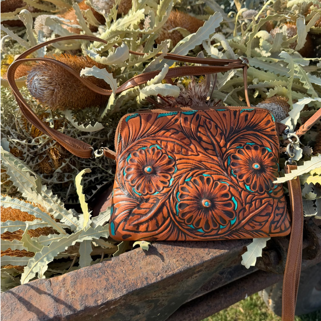 A brown tooled leather crossbody bag with turquoise accents, resting on a rustic stone surrounded by green leaves and brown seed pods.