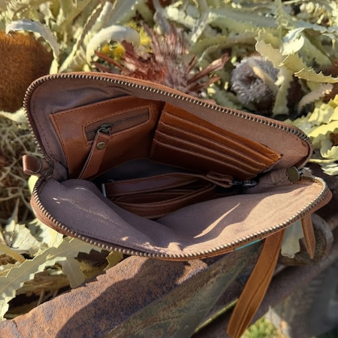 An open brown leather wallet or crossbody bag with multiple card slots, a zip pocket, and a long strap, photographed outdoors among dried greenery and a rusty metal surface.