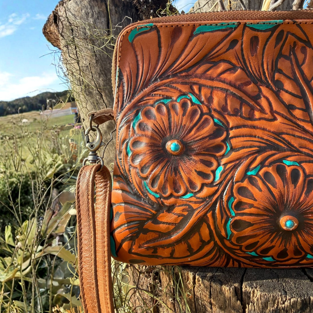 A tooled leather wristlet clutch with floral embossing and turquoise accents, shown outdoors on a rustic wooden post with a grassy field and trees in the background.