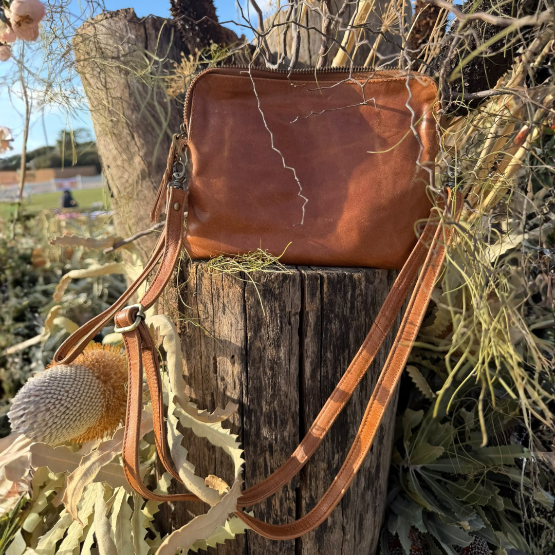 A brown leather crossbody bag with a long strap, displayed outdoors on a weathered wooden stump surrounded by dried plants and branches.