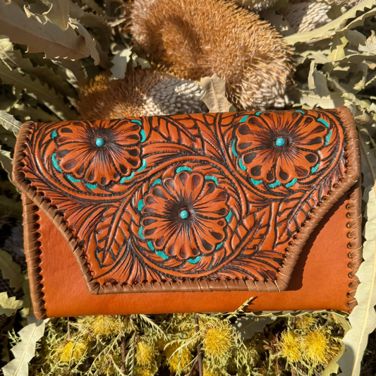 A hand-tooled leather clutch bag with intricate floral designs and turquoise accents, resting on a bed of dried leaves and seed pods.