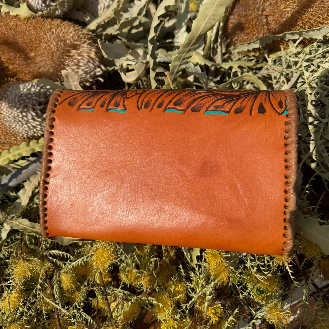 A handmade brown leather wallet with hand-stitched edges and a black hand-drawn tribal-style pattern across the top, resting on a bed of dried green and yellow plants.