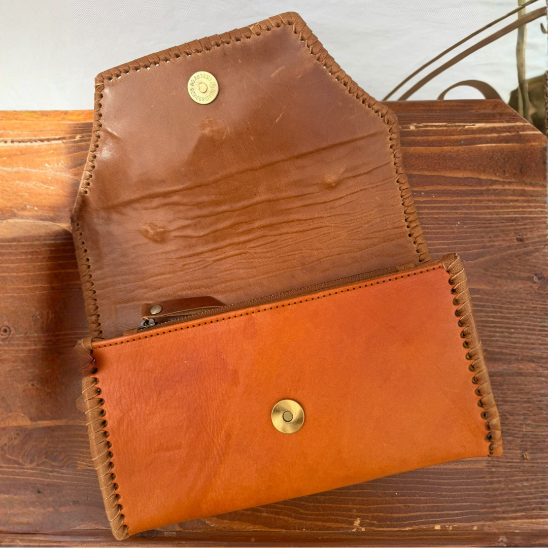 A handmade leather wallet in a warm cognac orange-brown color, shown open to reveal its interior. The wallet features a snap-button closure and a zippered pocket, with visible whipstitching along the edges, resting on a rustic wooden surface.
