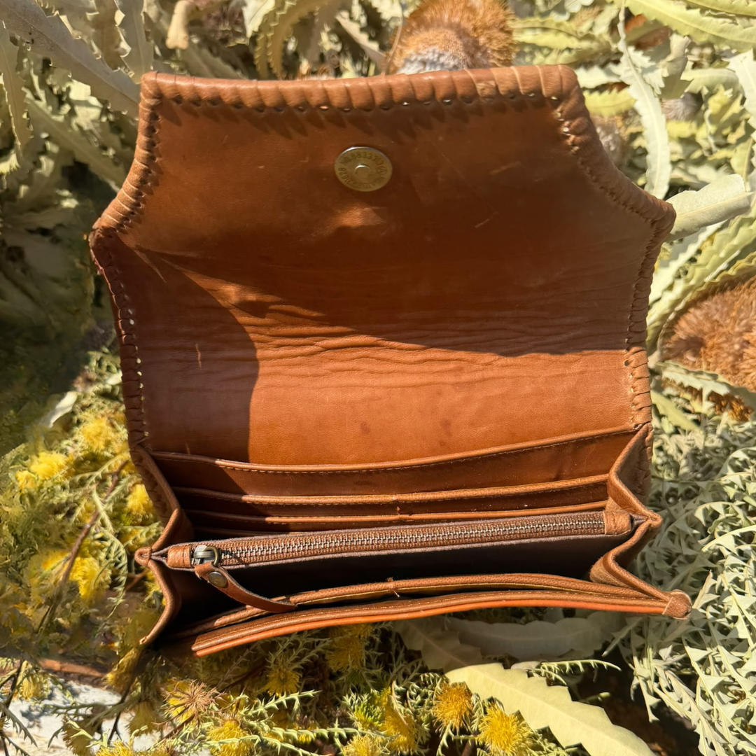 An open brown leather wallet or clutch with multiple card slots, a zip pocket, and a snap button closure, photographed outdoors among dried yellow flowers and green foliage.