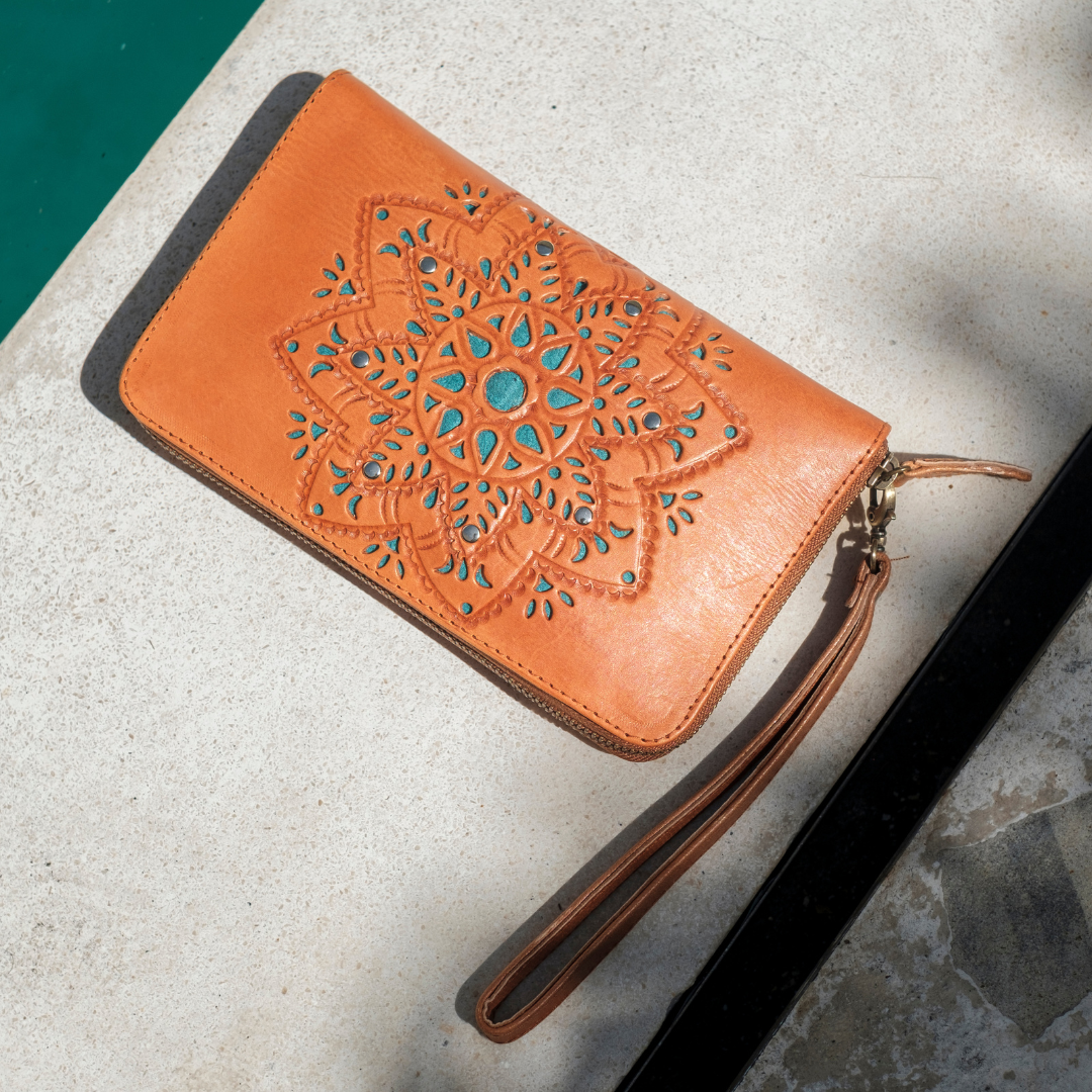 House of Hide Desert Mandala Wallet in orange leather with turquoise and brown mandala cutout design, wrist strap, and gold zipper being displayed on light gray stone