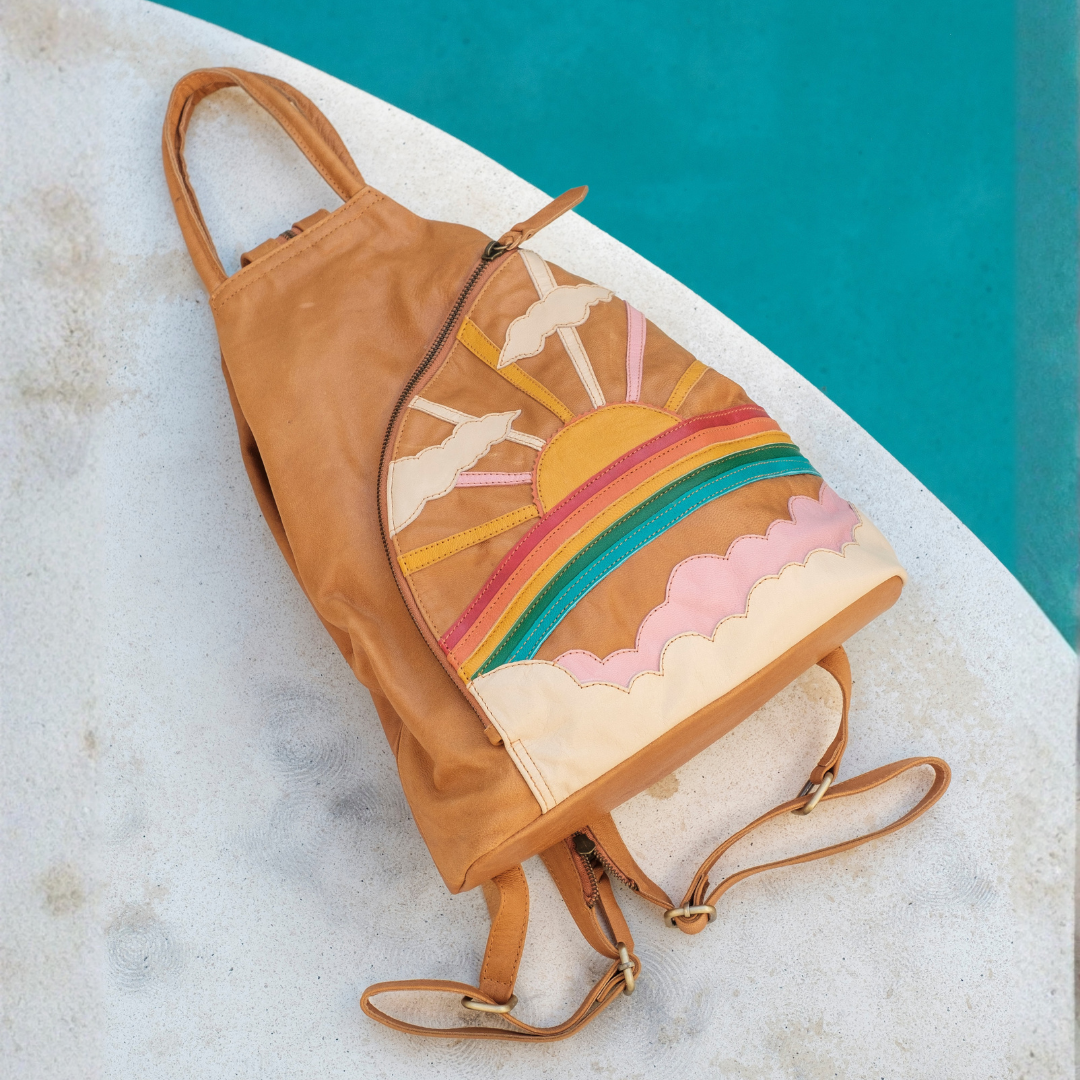 Desert Rainbow Backpack from House of Hide in tan leather with colorful patchwork design of sun, rainbow, clouds, and horse being displayed on white surface with turquoise background