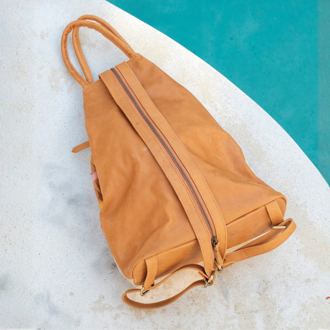 Desert Rainbow Backpack from House of Hide in tan leather with long shoulder strap and top handles being displayed on curved white surface with bright turquoise background