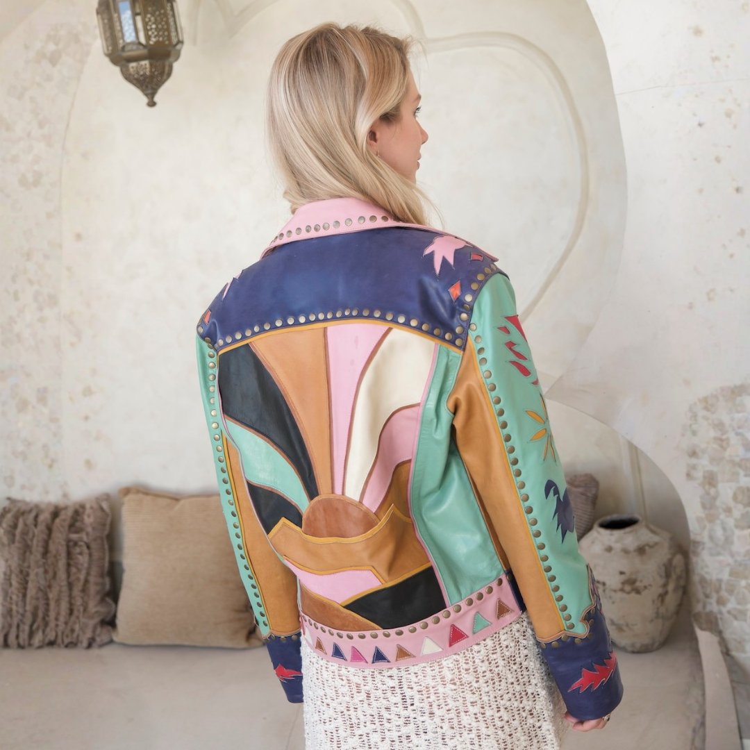 A woman viewed from behind wearing a colorful patchwork leather jacket with geometric panels in navy, mint green, pink, tan, and cream, set against a rustic, warm-toned interior background.