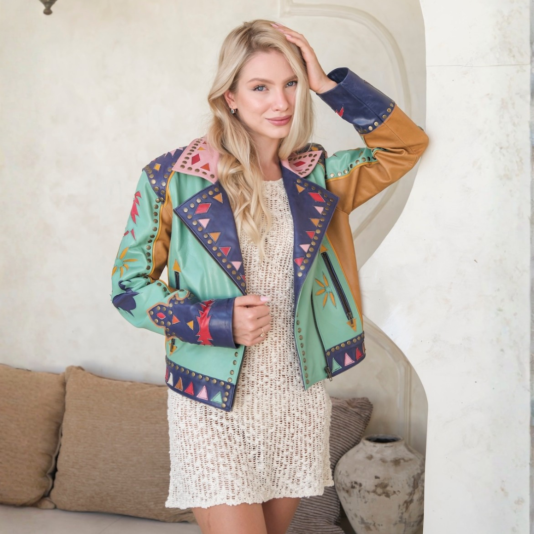 A blonde woman models a colorful patchwork moto jacket with green, navy, and tan panels, styled over a cream knit dress in a warm, neutral-toned interior setting.