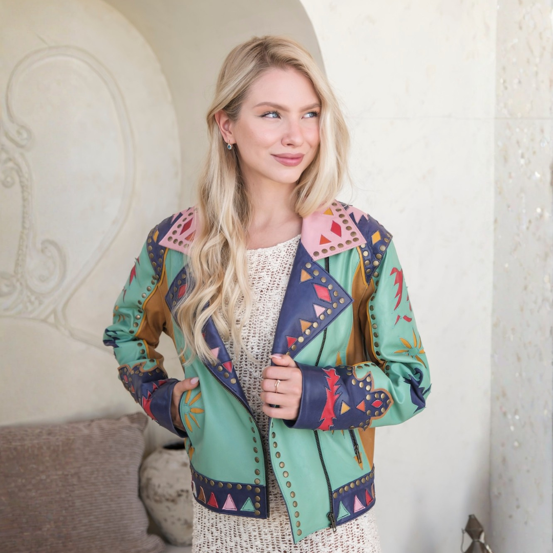 A blonde woman wearing a colorful patchwork leather jacket with Southwestern-style geometric patterns in mint green, navy blue, pink, and brown, styled over a cream knit top.
