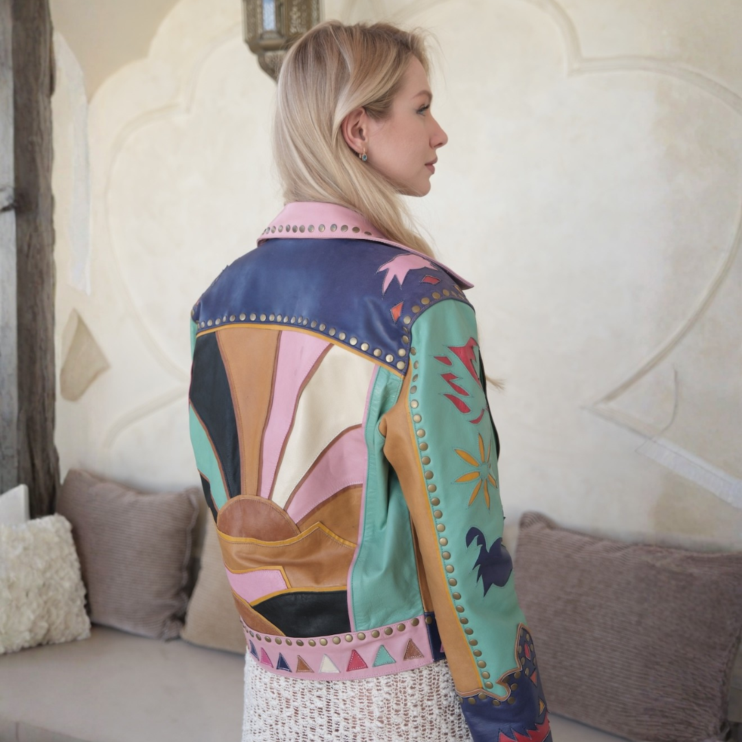 A woman shown from the back wearing a colorful patchwork leather jacket with geometric and floral patterns, set in a warm, rustic indoor environment.