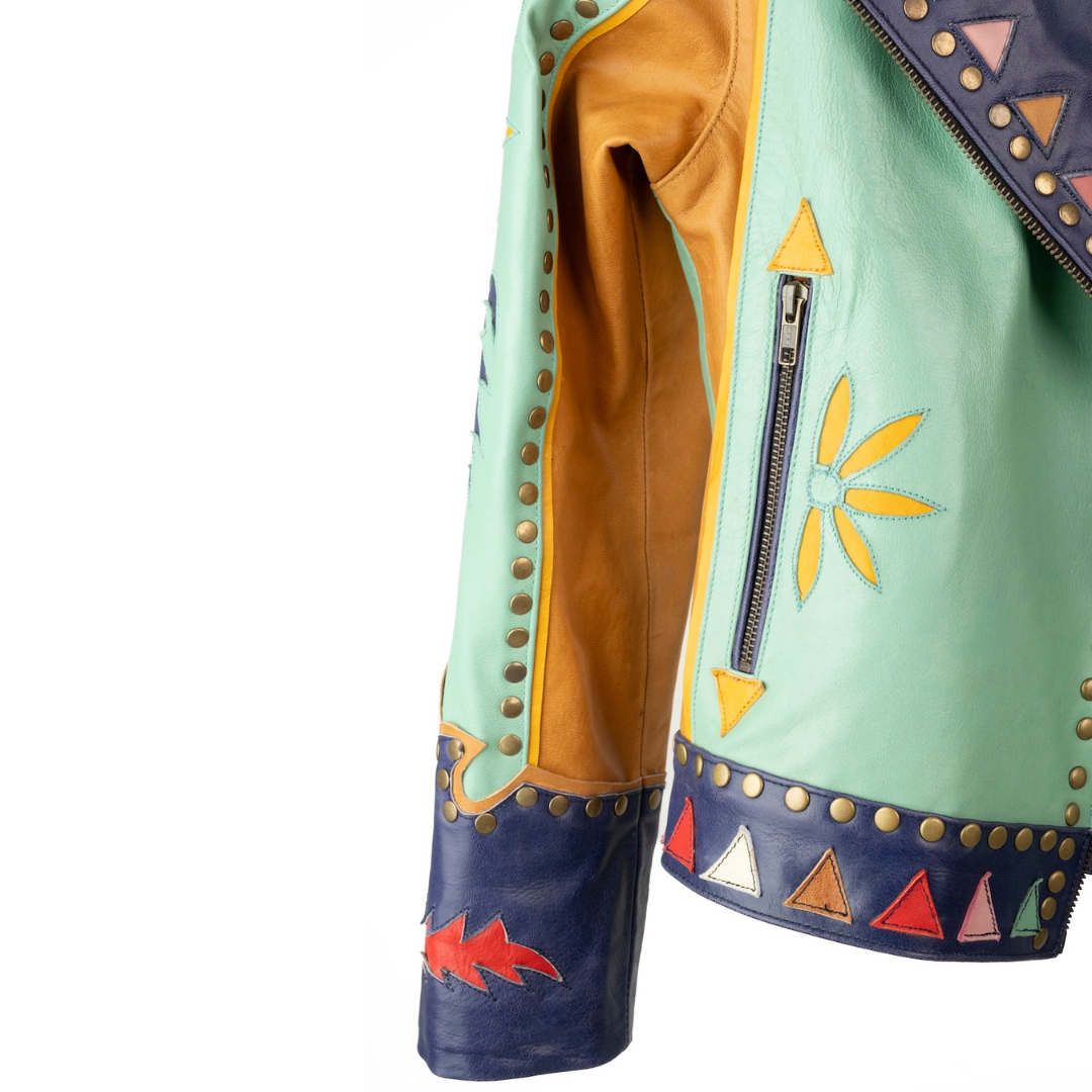 Close-up of a western-style leather jacket with mint green, tan, and navy blue panels, brass studs, and embroidered yellow and red details on a white background.
