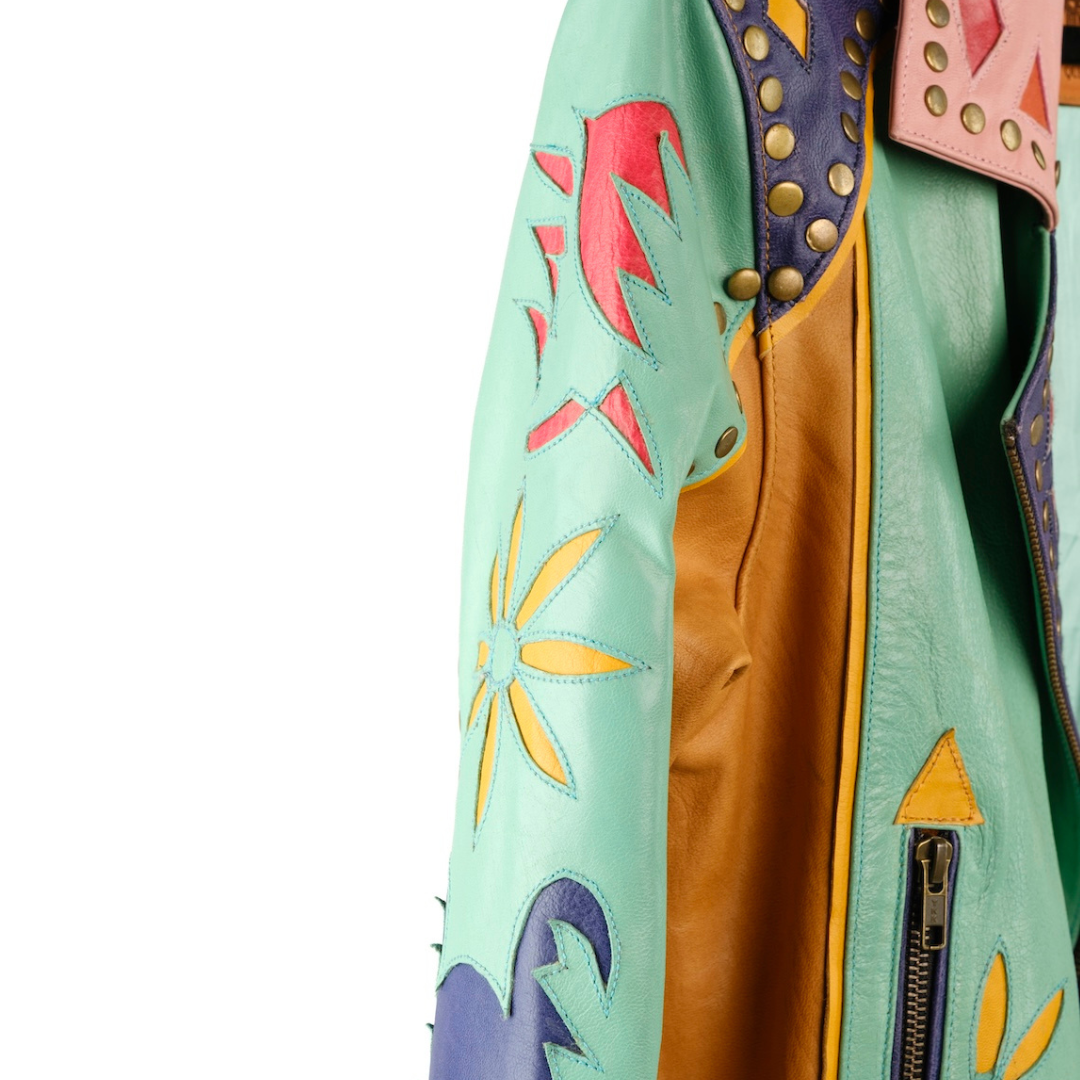 Close-up of a handcrafted leather jacket featuring a mint green body, a tan leather interior lining, purple shoulder panels with brass studs, and colorful embroidered floral motifs in pink and yellow.