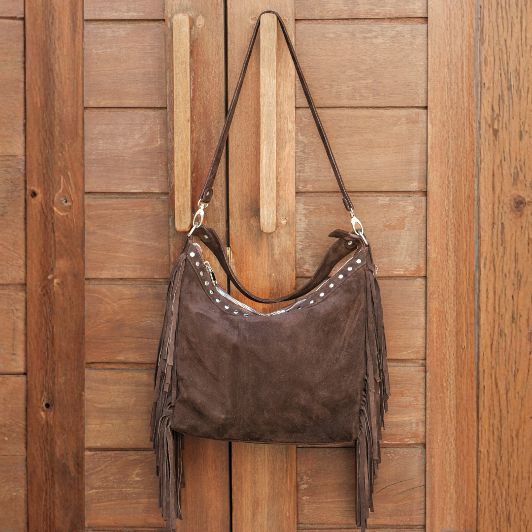 Drifter Fringe Bag from House of Hide in dark brown suede with fringe detail and silver studs being displayed hanging on wooden peg against rustic wooden door