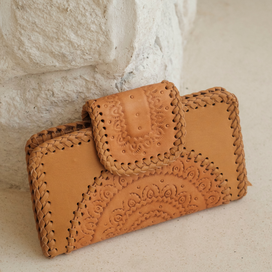 Dustwood Carved Wallet from House of Hide in tan leather with hand-tooled floral patterns and braided edges being displayed against textured white stone wall