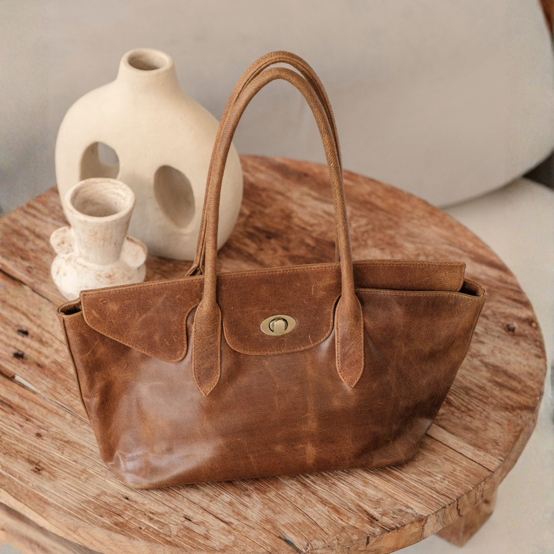 Frontier Tote Bag from House of Hide in brown leather with double top handles and snap-button closure being displayed on rustic wooden table with ceramic vases