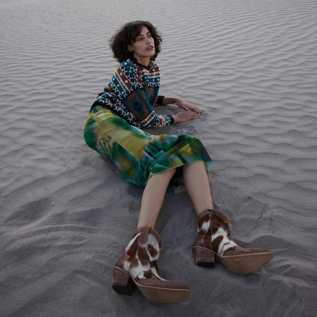 Model reclining on sand wearing Georgia tan and white cowhide ankle boots with tooled leather and block heels.
