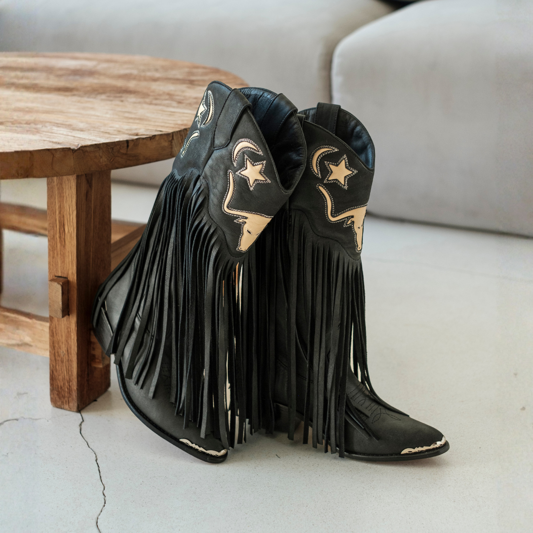 A pair of black leather fringe cowboy boots with gold-toned western embroidery, resting on a light concrete floor next to a wooden table.