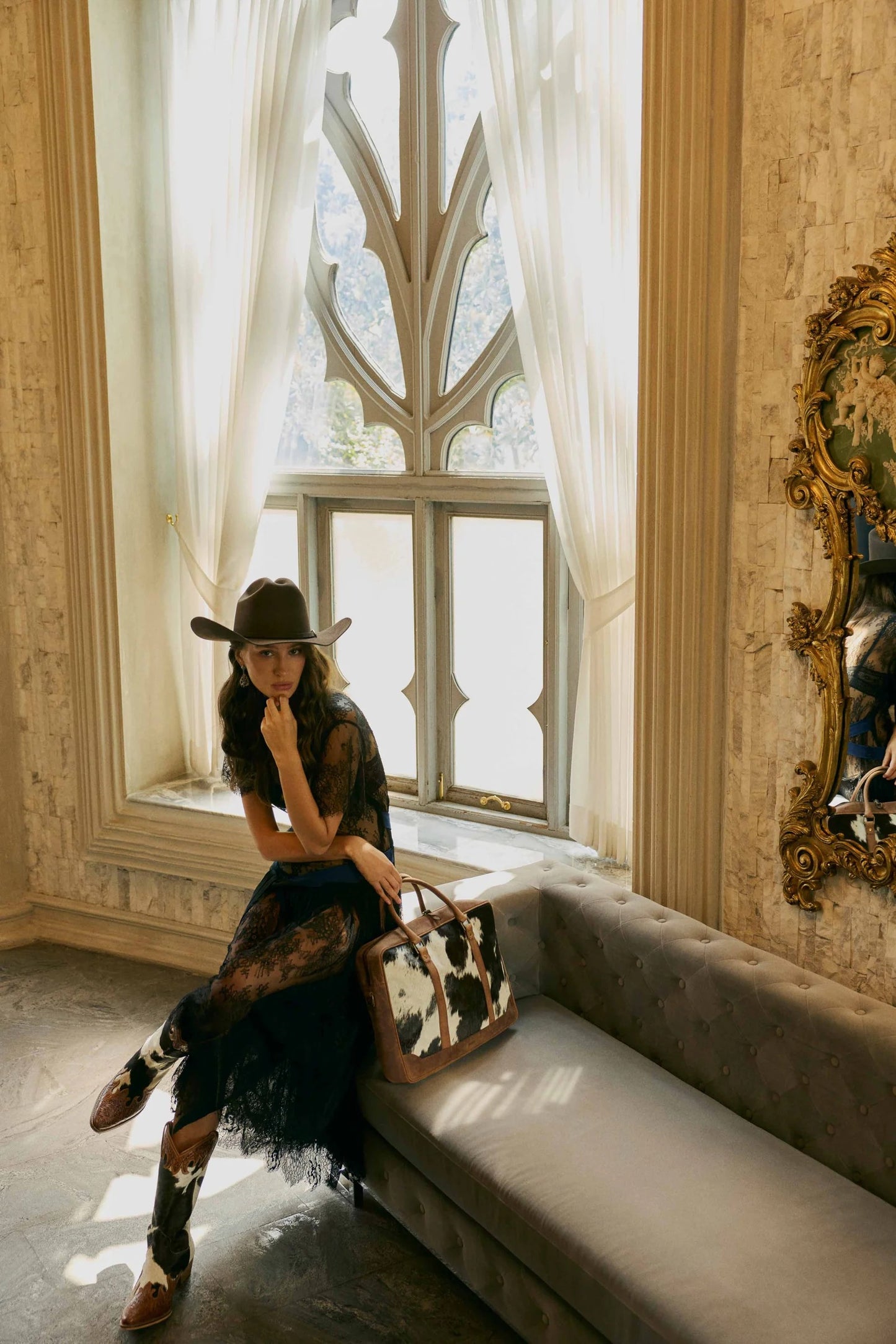Model showcasing House of Hide women's western fashion while seated on a couch in an elegant room with large windows and refined decorative details