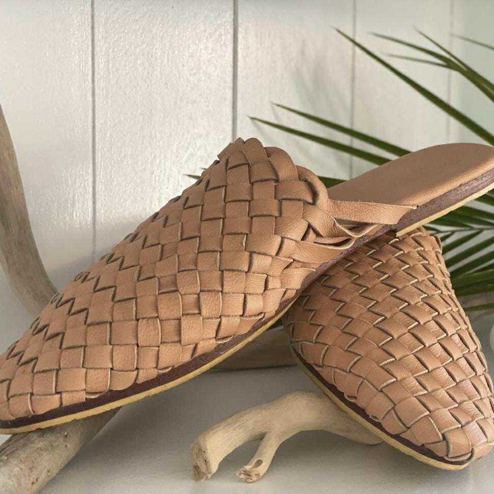 Pair of woven brown mules on a light wooden surface with a plant in the background