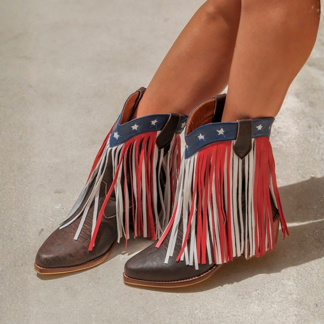 A pair of women's western-style ankle boots with long fringe in red, white, and brown, featuring a blue leather strap with white stars at the collar, shown on a model's feet against a concrete background.